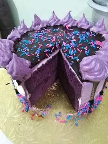 A delicate  Purple Vanilla cake with purple vanilla frosting, a rich chocolate drip, and pink, purple, and blue sprinkles to accent the top and bottom sides of the cake.